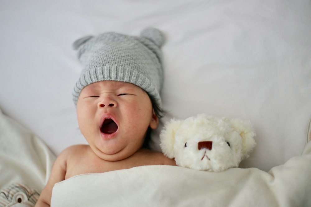 Un bébé qui s'endort, un bonnet gris sur la tête, à côté d'un petit ours en peluche